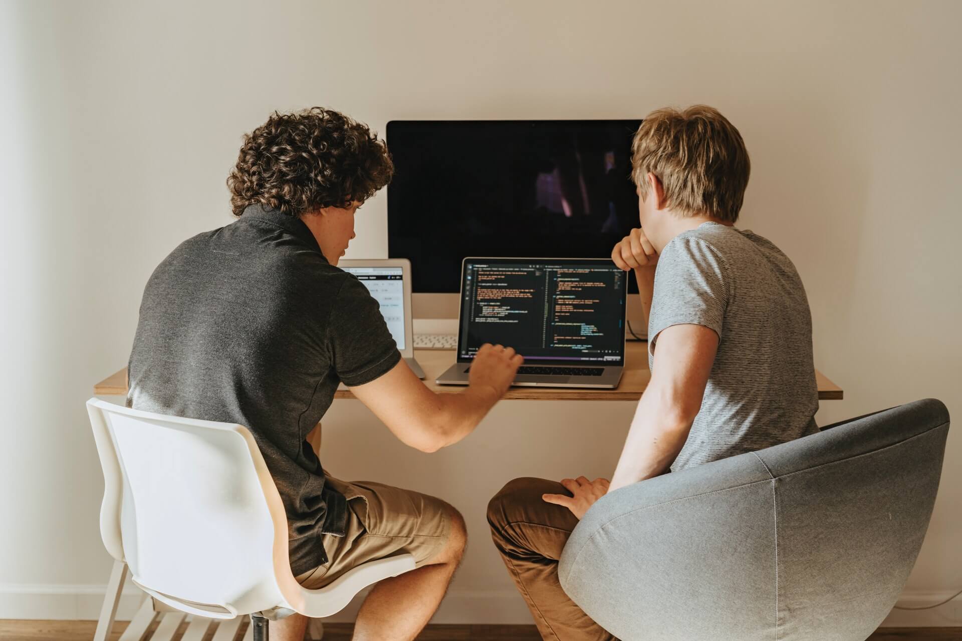 Two person working on their laptop working some code with a big monitor display upfront on top the table, showcasing a modern setup in web development.